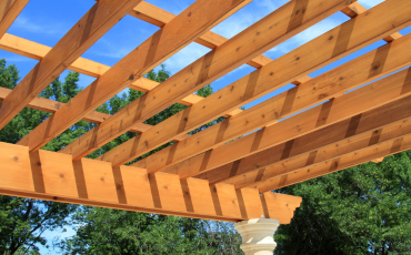 A photo of the top part of a pergola structure. You can see a blue sky and green trees in the background.