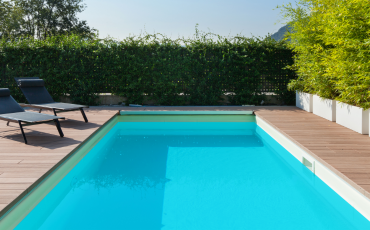 A photo of a luxurious pool with a beautiful and simplistic pool deck surrounding the pool. The pool has two pool chairs on the deck to the left of the pool. There is also a fence of green shrubs acting as a fence around the back yard.