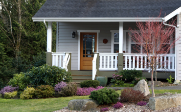 This is a photo of a porch in front of a small house. The porch is covered with a roof. The porch is also surrounded by green shrubbery and bushes. It is a beautiful photo of a beautiful house.