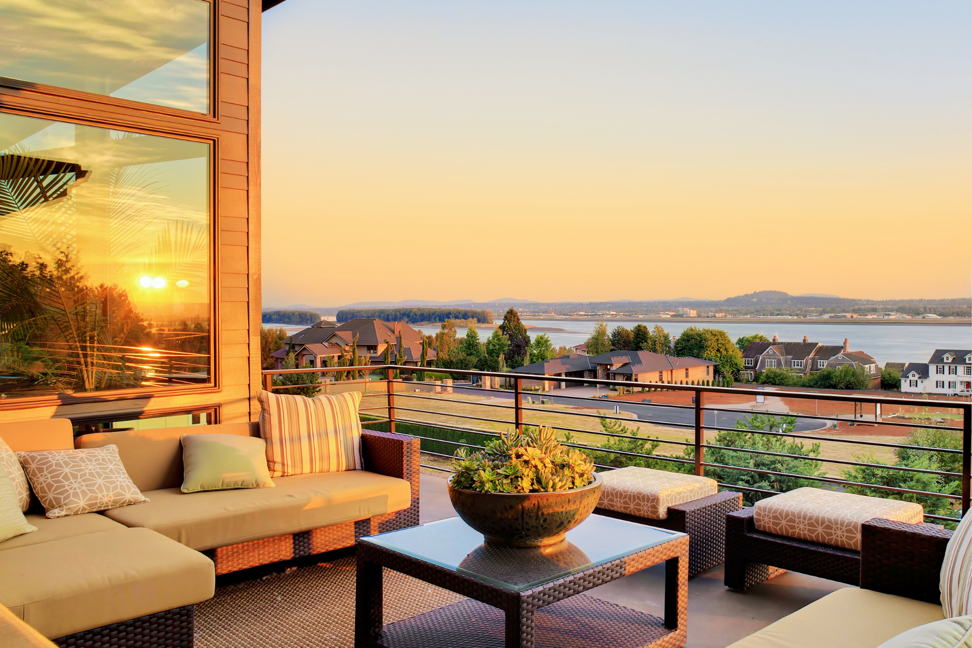 A photo of a roof deck on the roof of a building. The deck has plenty of furniture to make it look fun and cozy. The sun is starting to set and you can see the sun setting in the reflection of the glass on the left of the photo.