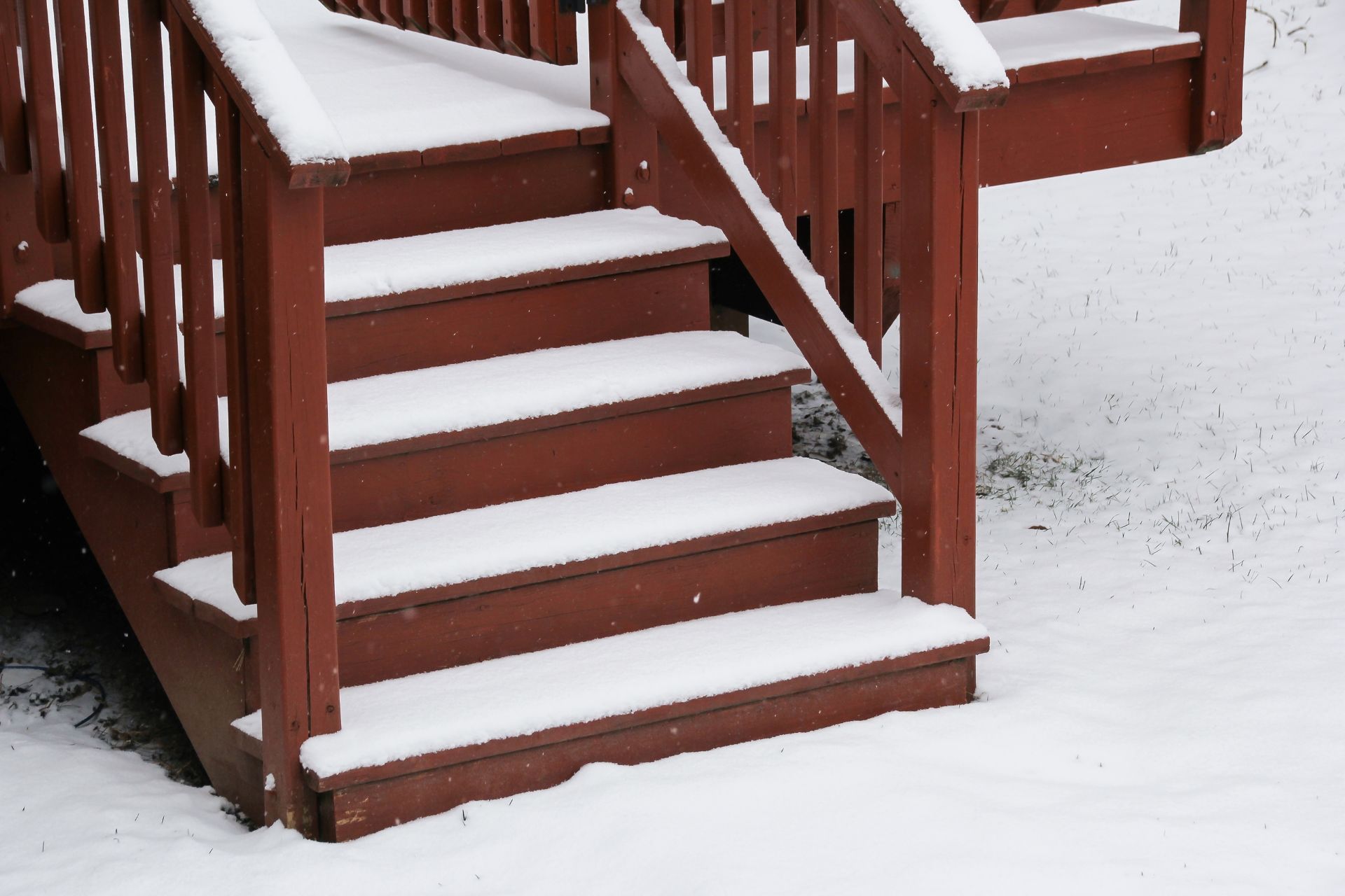 Snow-covered wooden stairs lead down from a deck, with each step accumulating a pristine layer of snow in St. Paul, Minnesota. The contrasting dark brown paint of the wood and the white snow create a peaceful winter scene. The deck's railing is also dusted with snow, and snowflakes are scattered across the surrounding ground, indicating a recent snowfall.