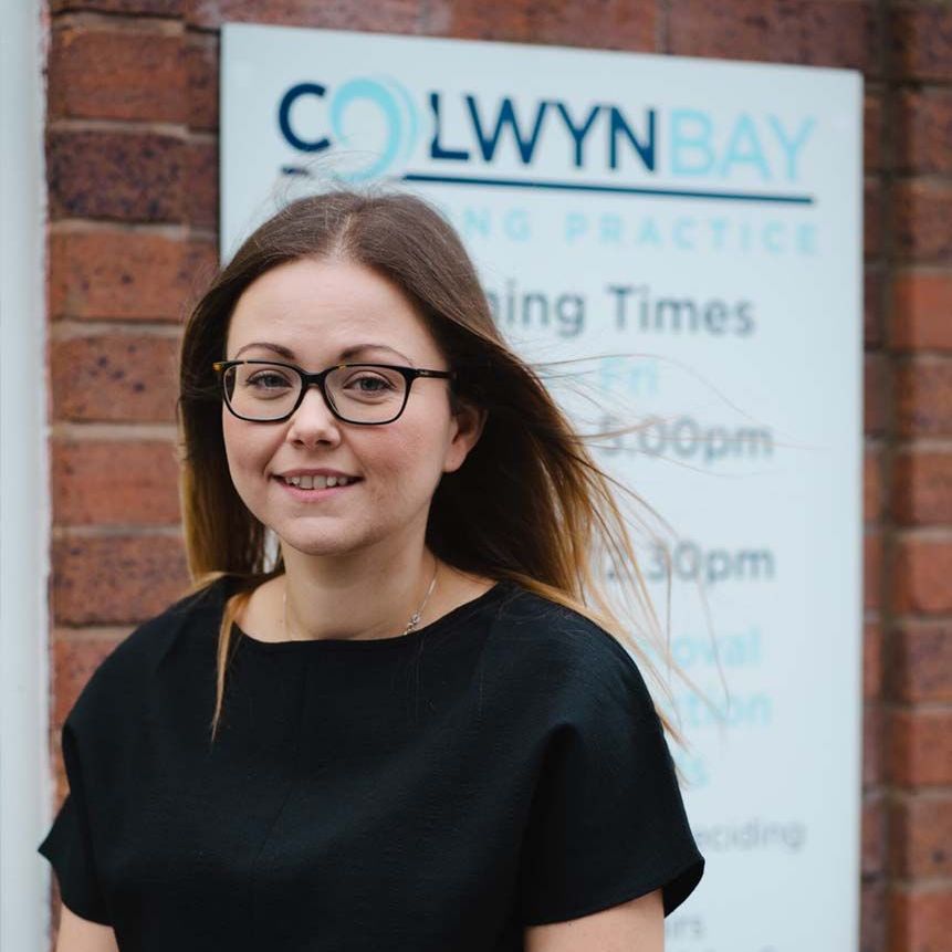 A woman wearing glasses is standing in front of a colwyn bay sign.