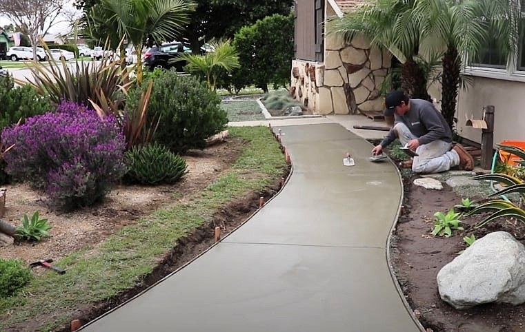 Worker finishing freshly poured concrete walkway in residential front yard landscaping.