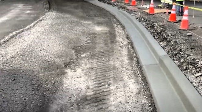 Fresh concrete curb beside an unfinished asphalt road with tire tracks and safety cones