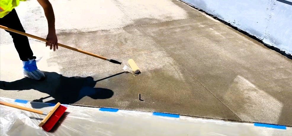 Person applying sealant to concrete with paint roller, protective sheeting on edges.