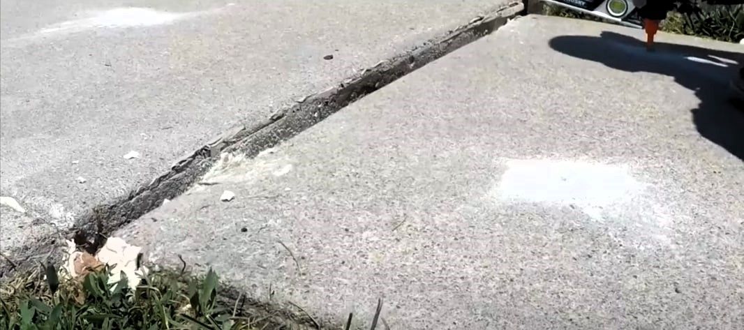Expert caulking a sidewalk crack in uneven concrete slabs, with grass growing at the edges.