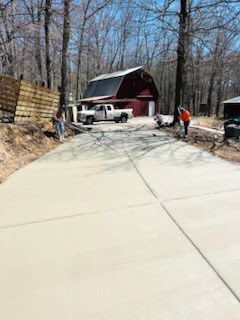 A concrete driveway leading to a red barn in the woods.