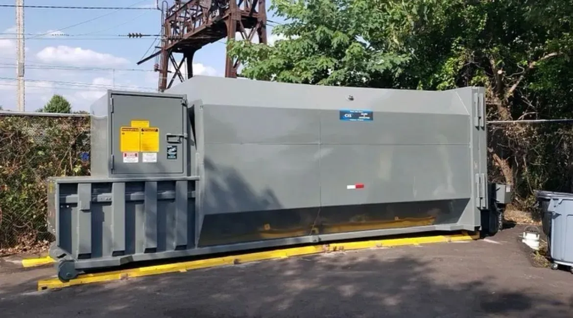 Large, gray commercial trash compactor near a fence and bushes, with safety signage on the side.
