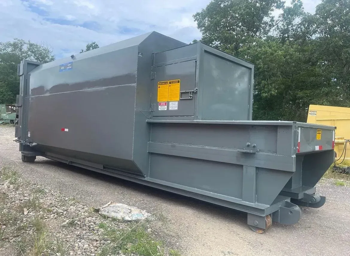 Gray industrial waste compactor trailer on a gravel surface.