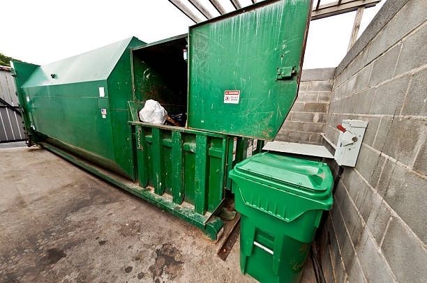 Green trash compactor with open door and trash can next to a cinder block wall.