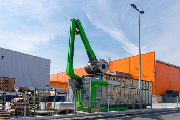 Green crane loading metal cylinder into a container. Industrial setting, orange and white buildings in background.