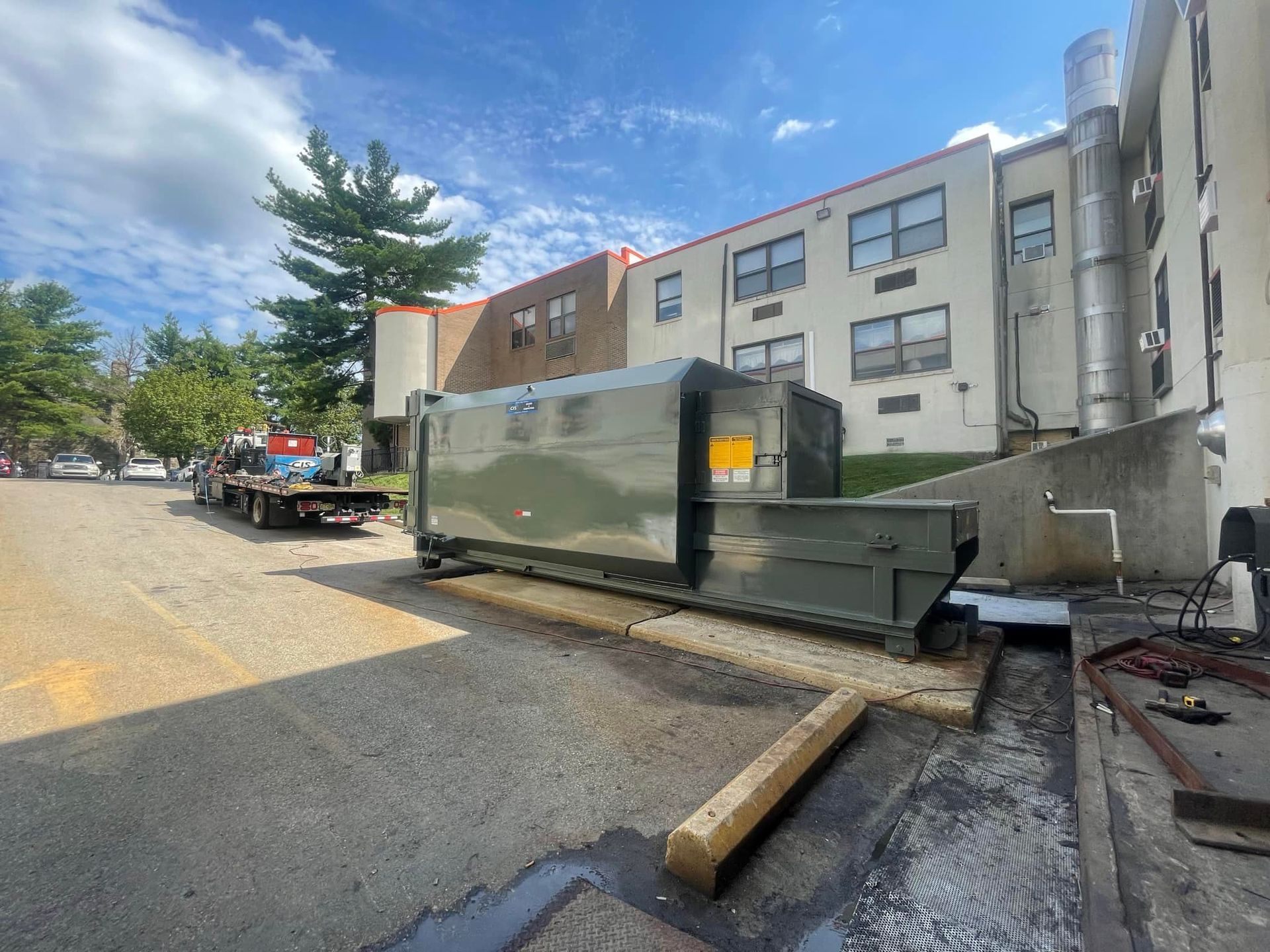 Large green dumpster on asphalt, next to a building. Flatbed truck in background.