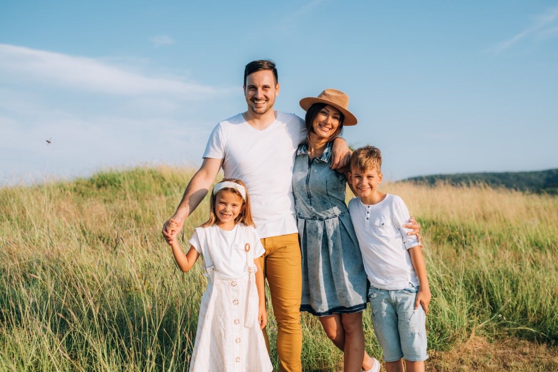 Family of four smiling together in a field of tall grass under a blue sky.