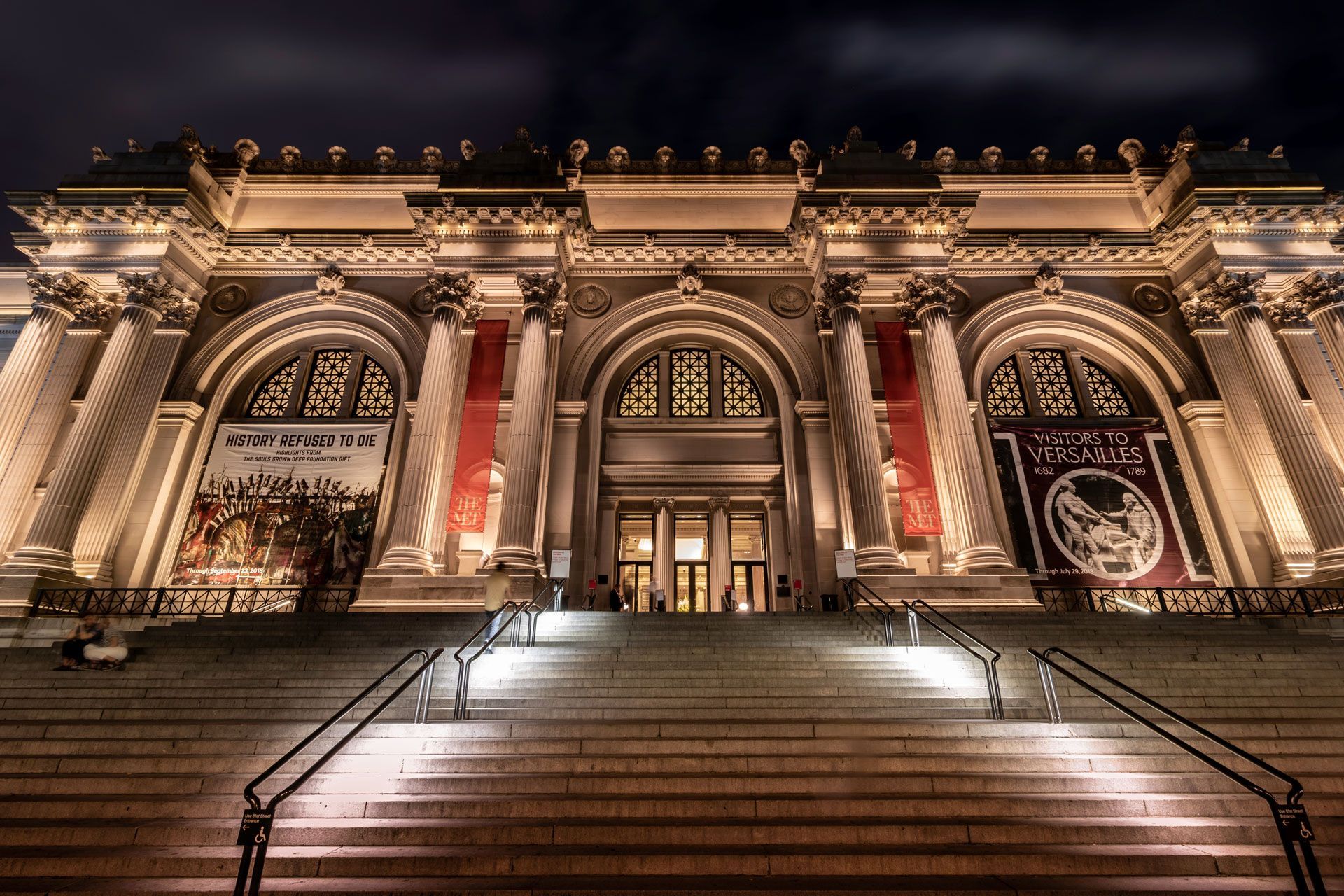 A large building with stairs leading up to it is lit up at night.