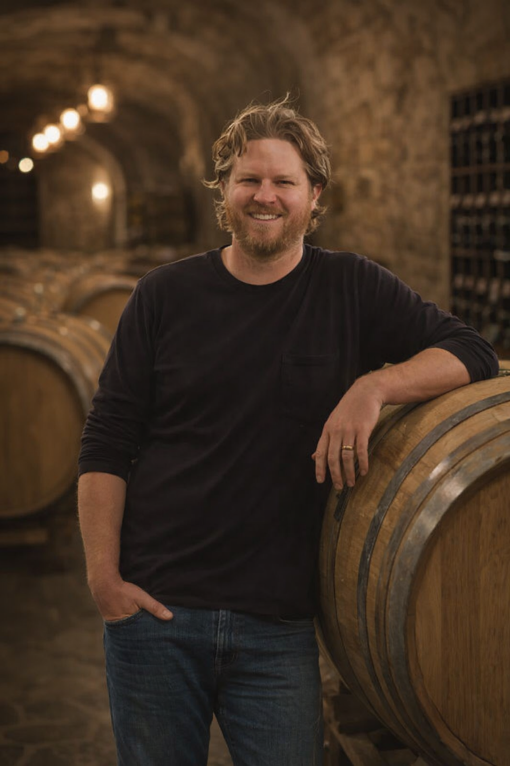 A man in a plaid shirt stands in front of a row of wine barrels