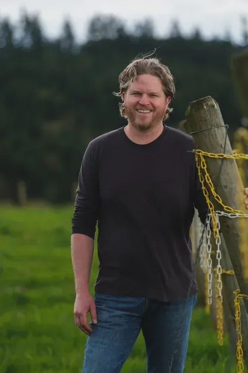 A man in a plaid shirt stands in front of a row of wine barrels