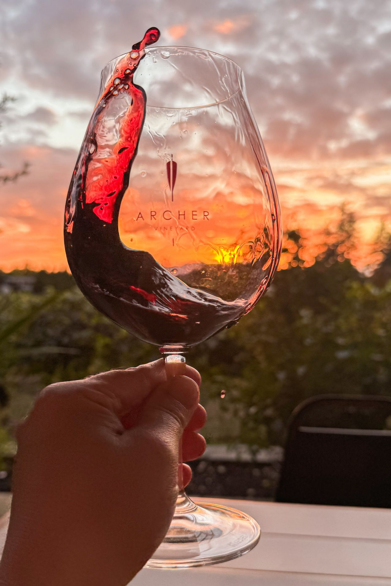 Hand holding a wine glass with red wine splashing, sunset in background.