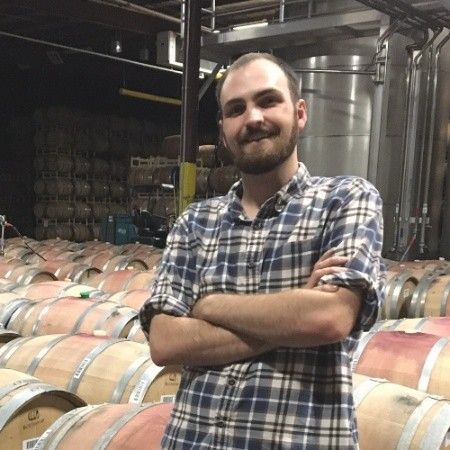 A man in a plaid shirt stands in front of a row of wine barrels