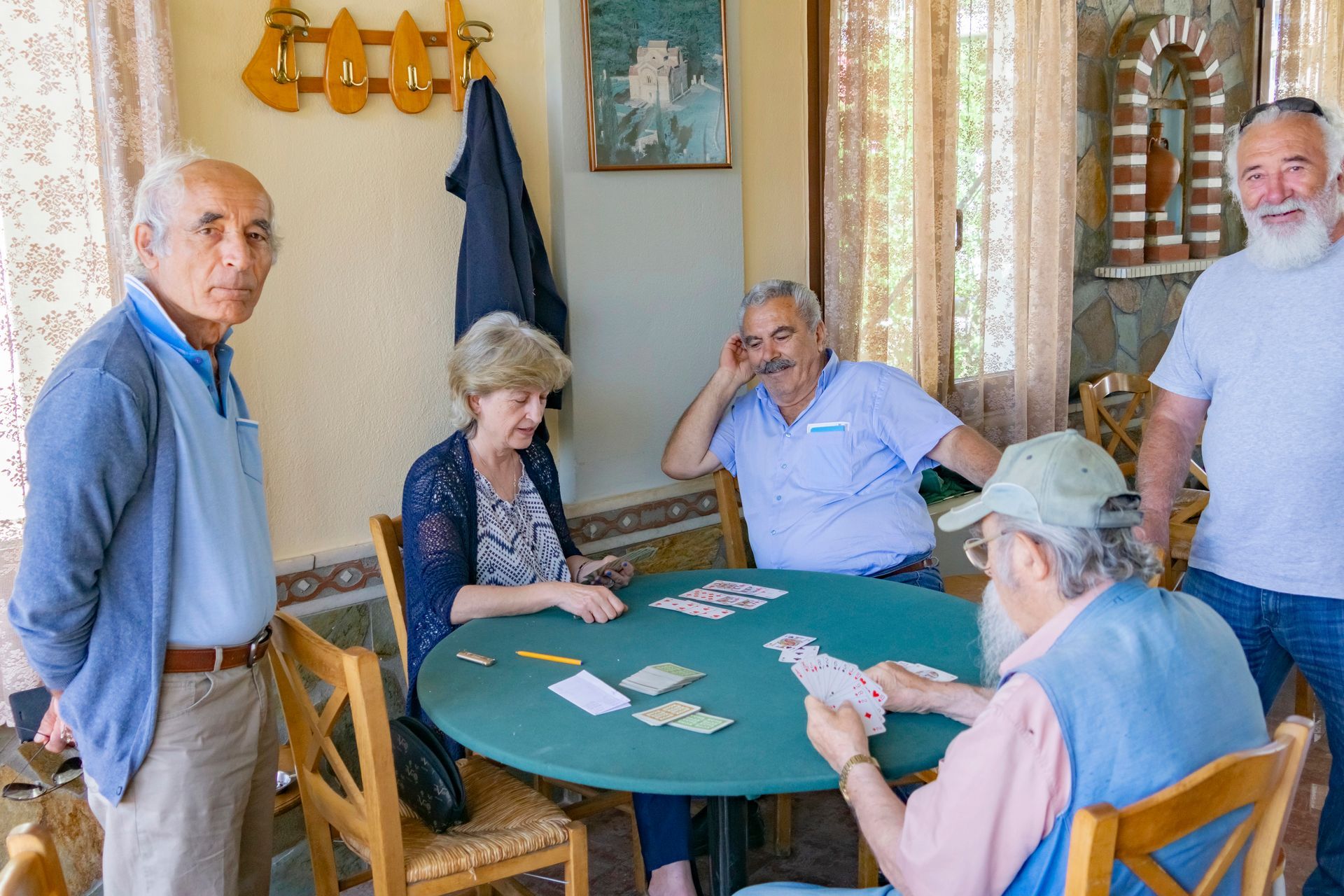 Five people playing cards around a green table indoors; one looks at the camera.