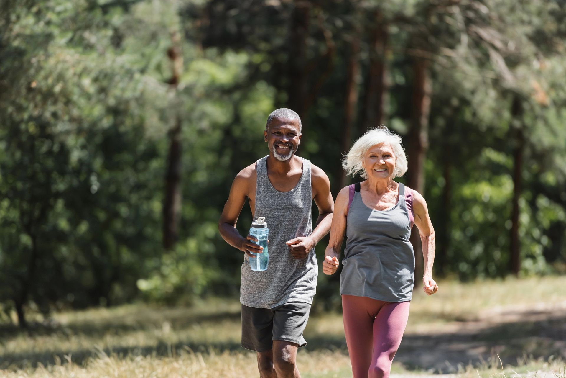 Two older adults running on a path in a park. The man holds a water bottle and smiles. The woman also smiles.