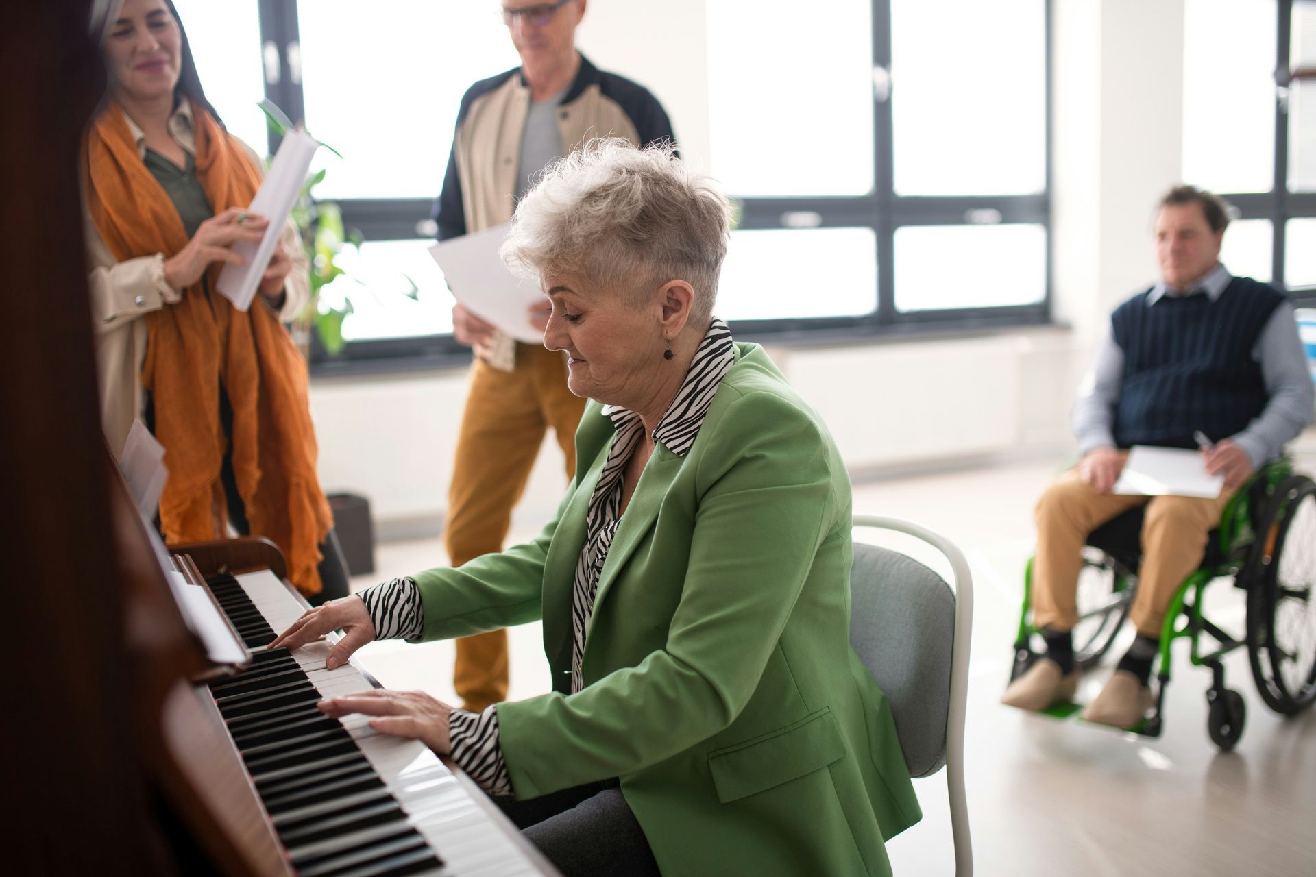 Elderly woman plays piano in a group music class. People sing, including a man in a wheelchair. Bright room.