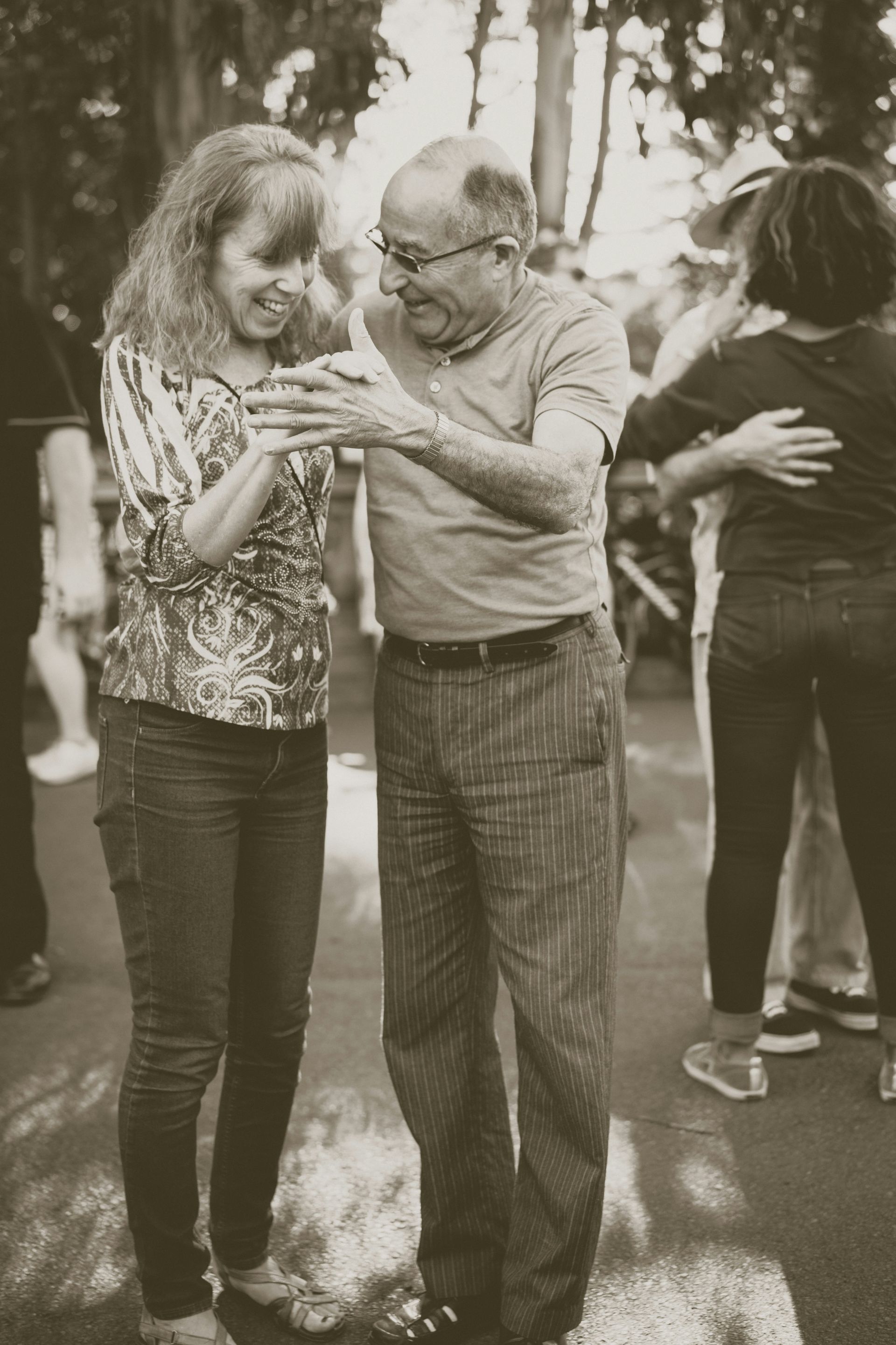 Woman and elderly man smiling and dancing outdoors. Other people dance in background.