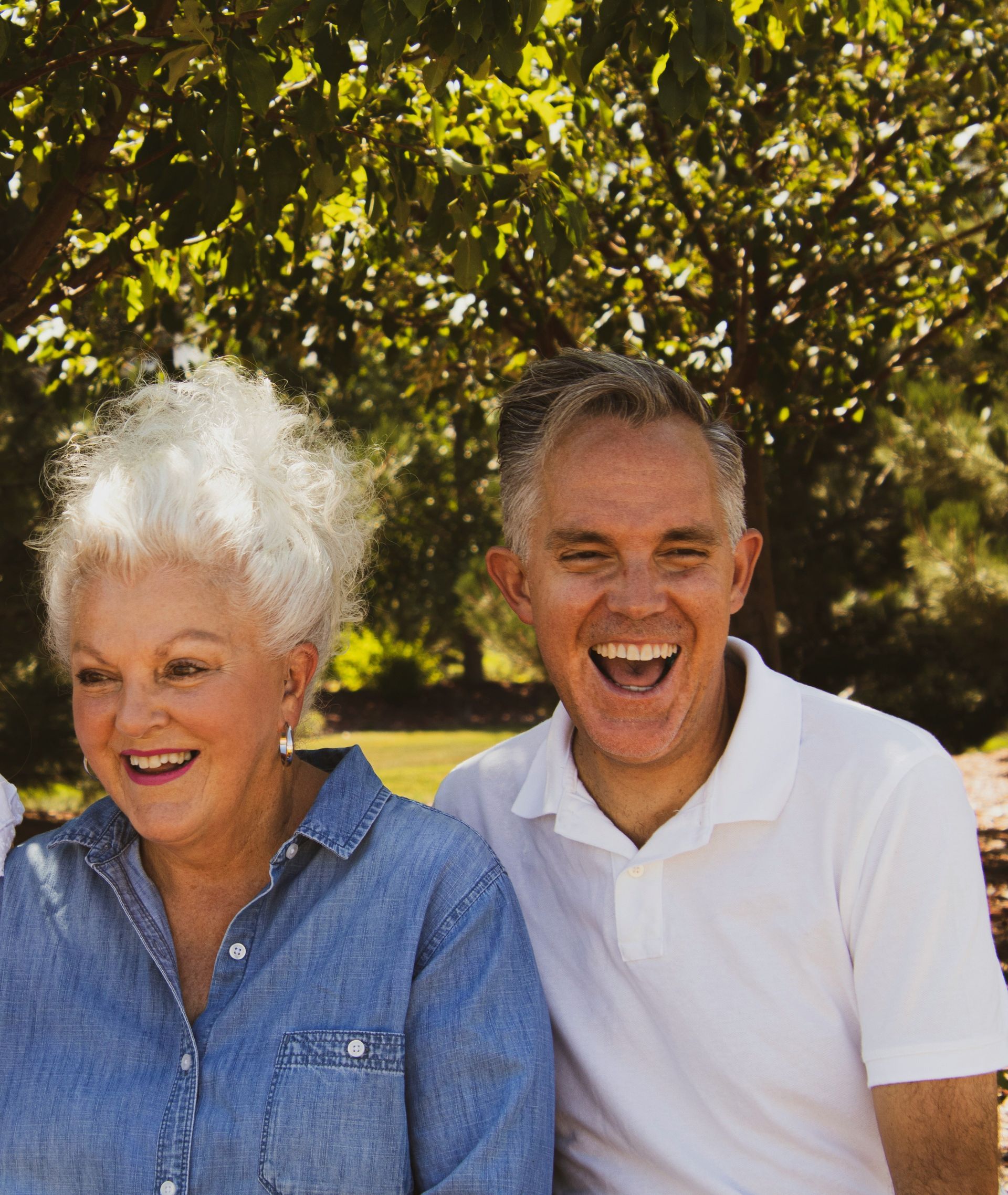 Woman and man laughing outdoors under trees; woman in denim shirt, man in white polo.