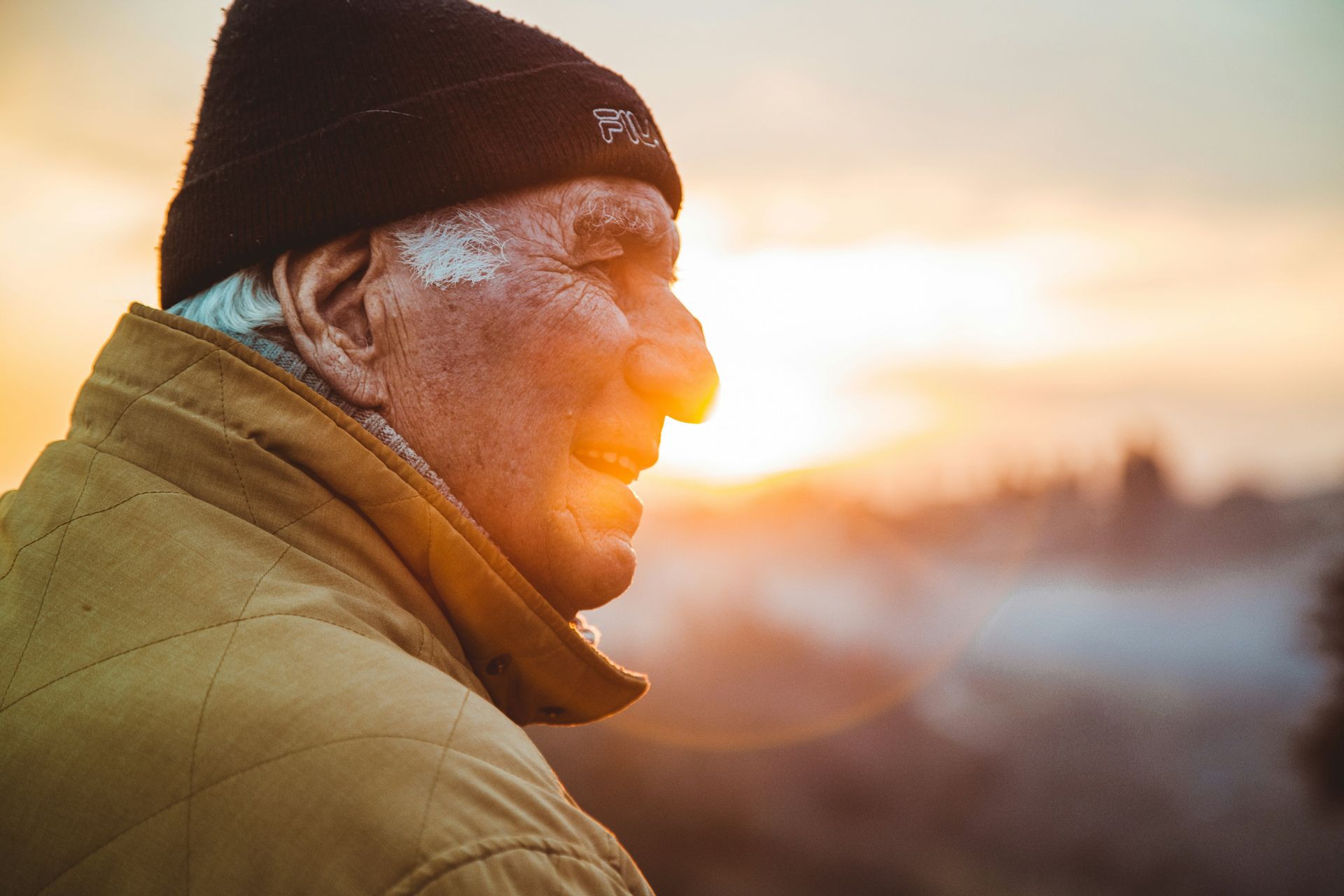Elderly man in a brown jacket and black hat, illuminated by a sunrise, looking off into the distance.