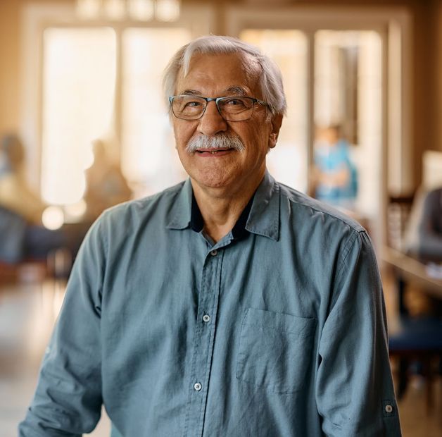 Elderly man with gray hair and mustache wearing glasses and blue shirt, smiles in a bright room.