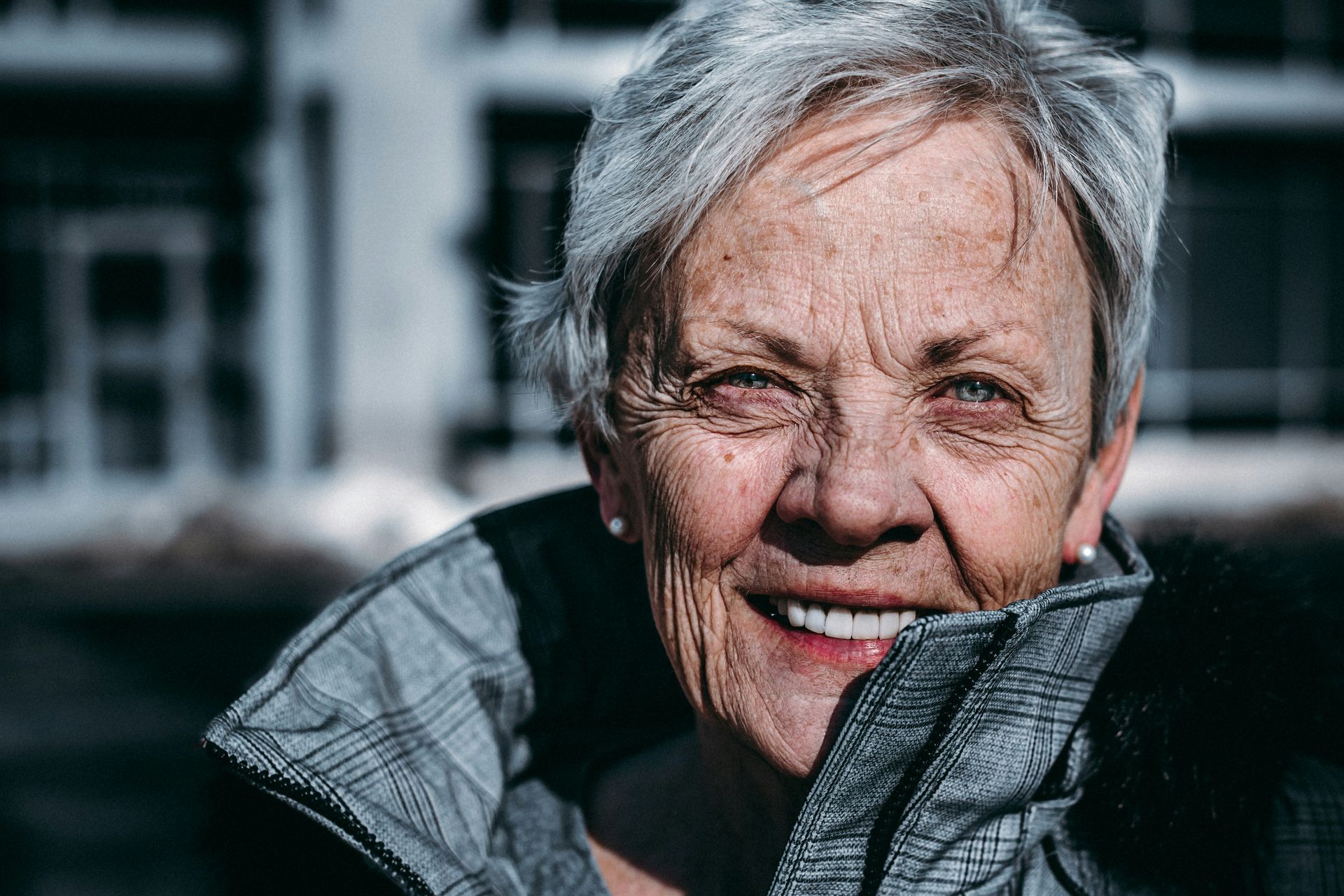 Senior woman with short gray hair smiles broadly outdoors, wearing a gray coat.