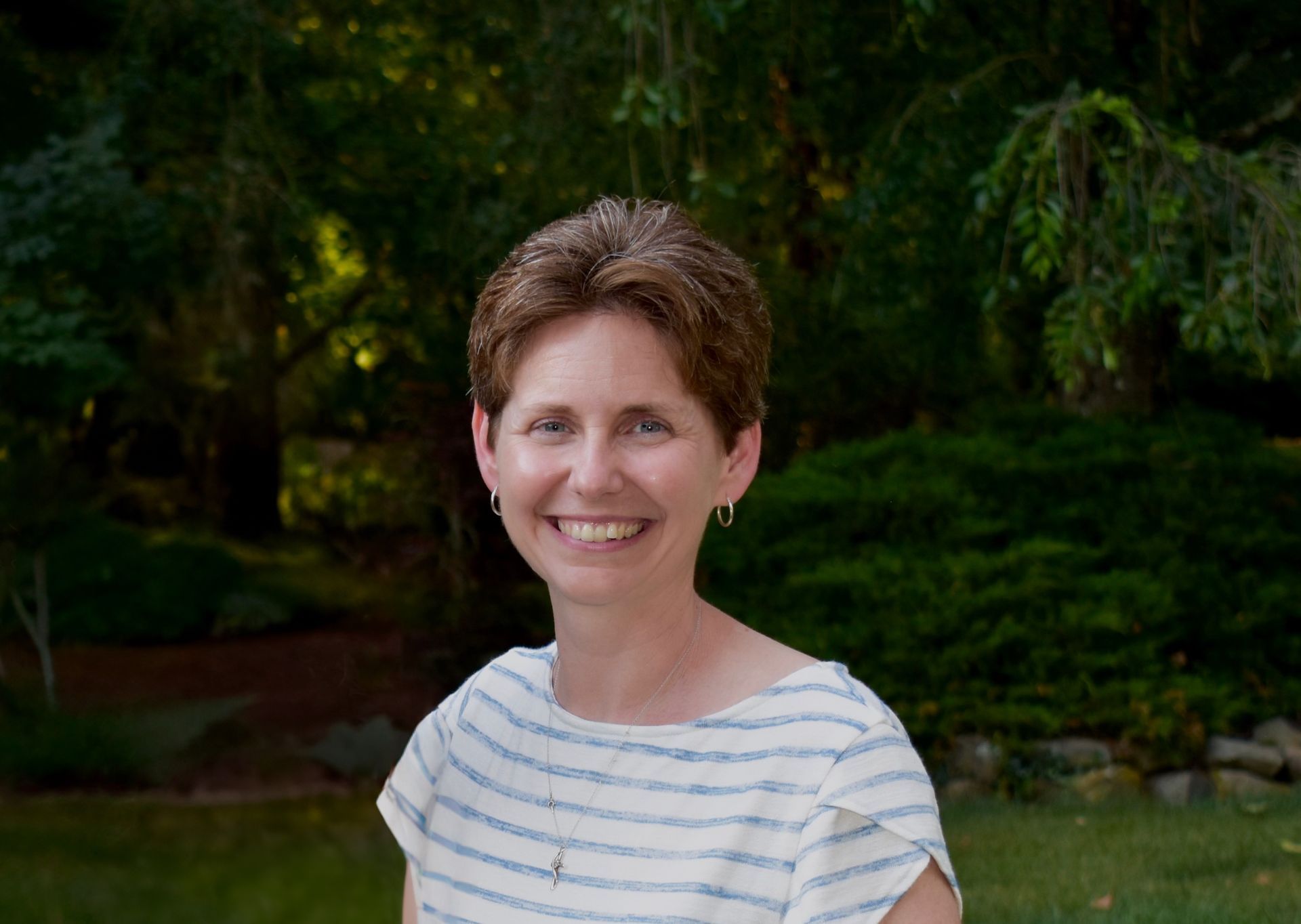 A woman in a striped shirt is smiling in front of trees.