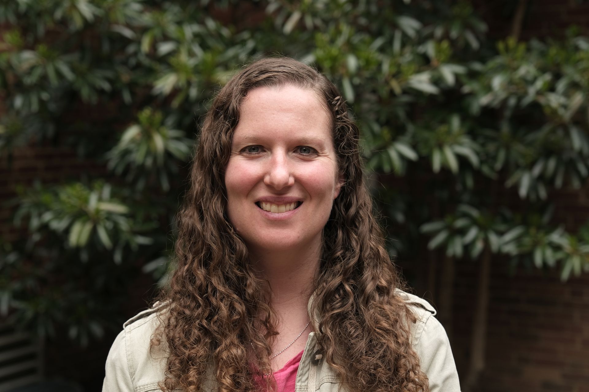 A woman with long curly hair is smiling in front of a tree.