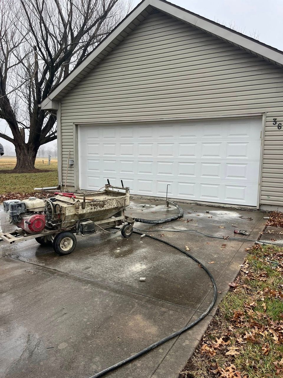 Concrete mixer cart outside a garage with a white door, hose on wet driveway, and fallen leaves