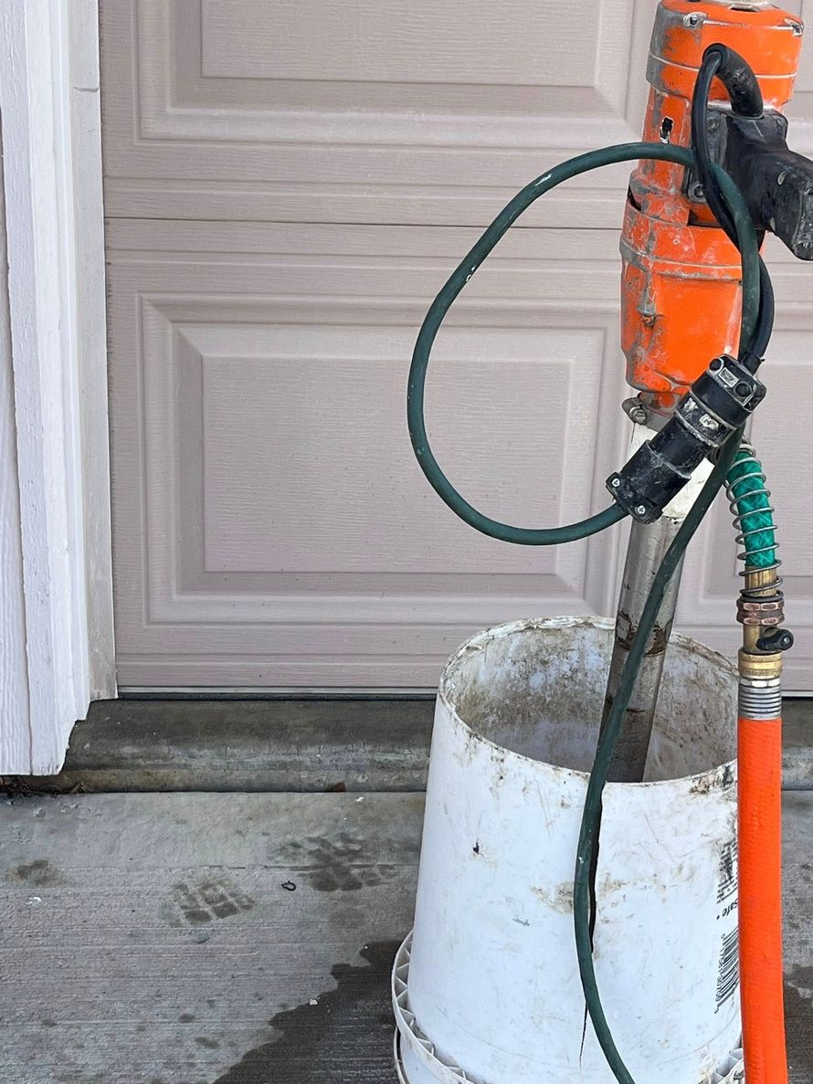 Orange pressure-washing machine beside a beige garage door, with hoses and a bucket on a concrete driveway.