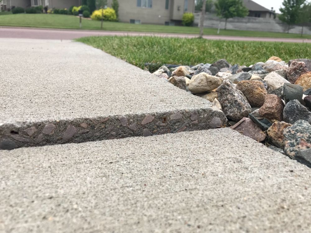 Cracked concrete sidewalk edge beside a decorative rock bed near a street and houses