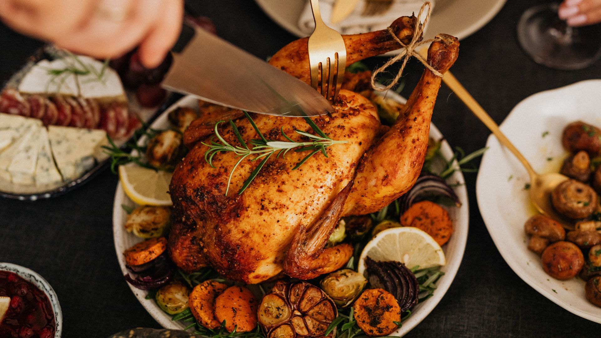 Roasted chicken being carved at a holiday table, surrounded by sides like cheese, potatoes, and mushrooms.