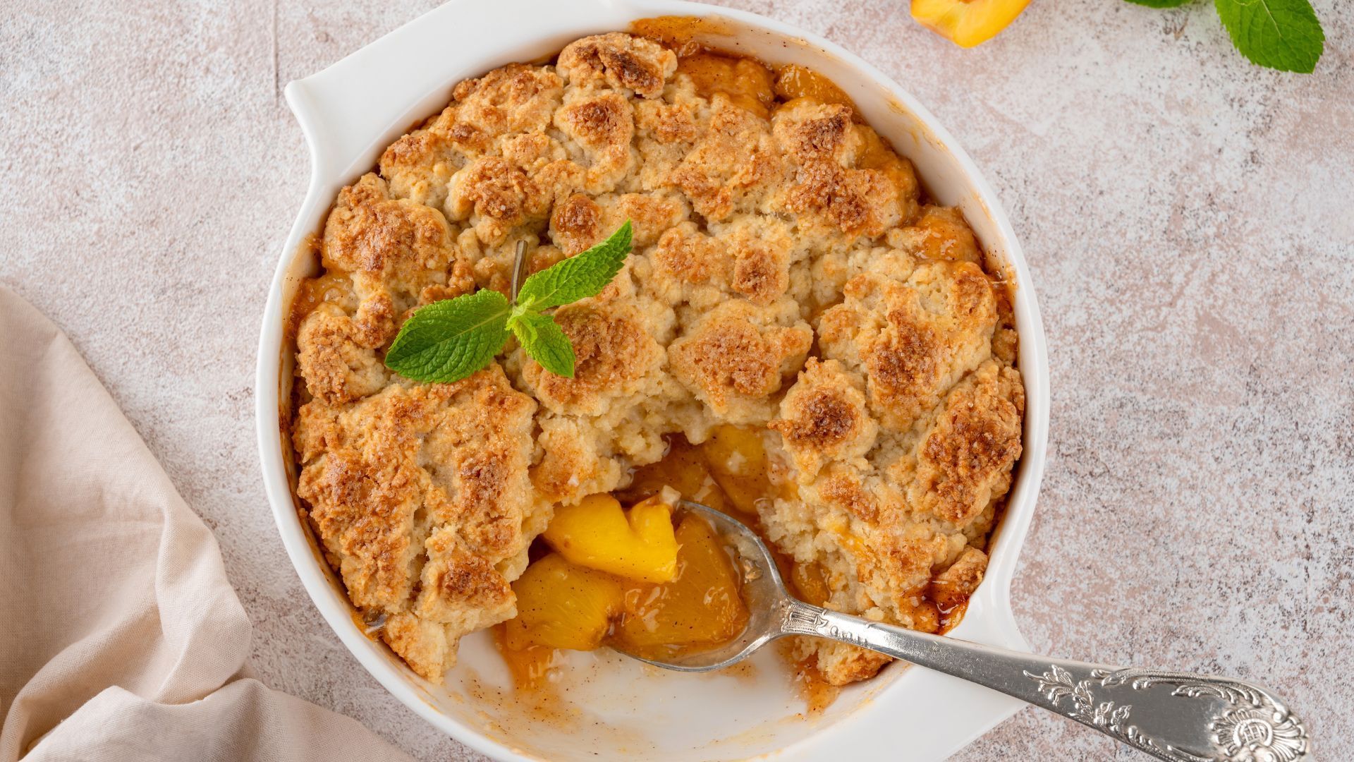 Peach cobbler in a white dish with a spoon; topped with golden crumble, garnished with mint.