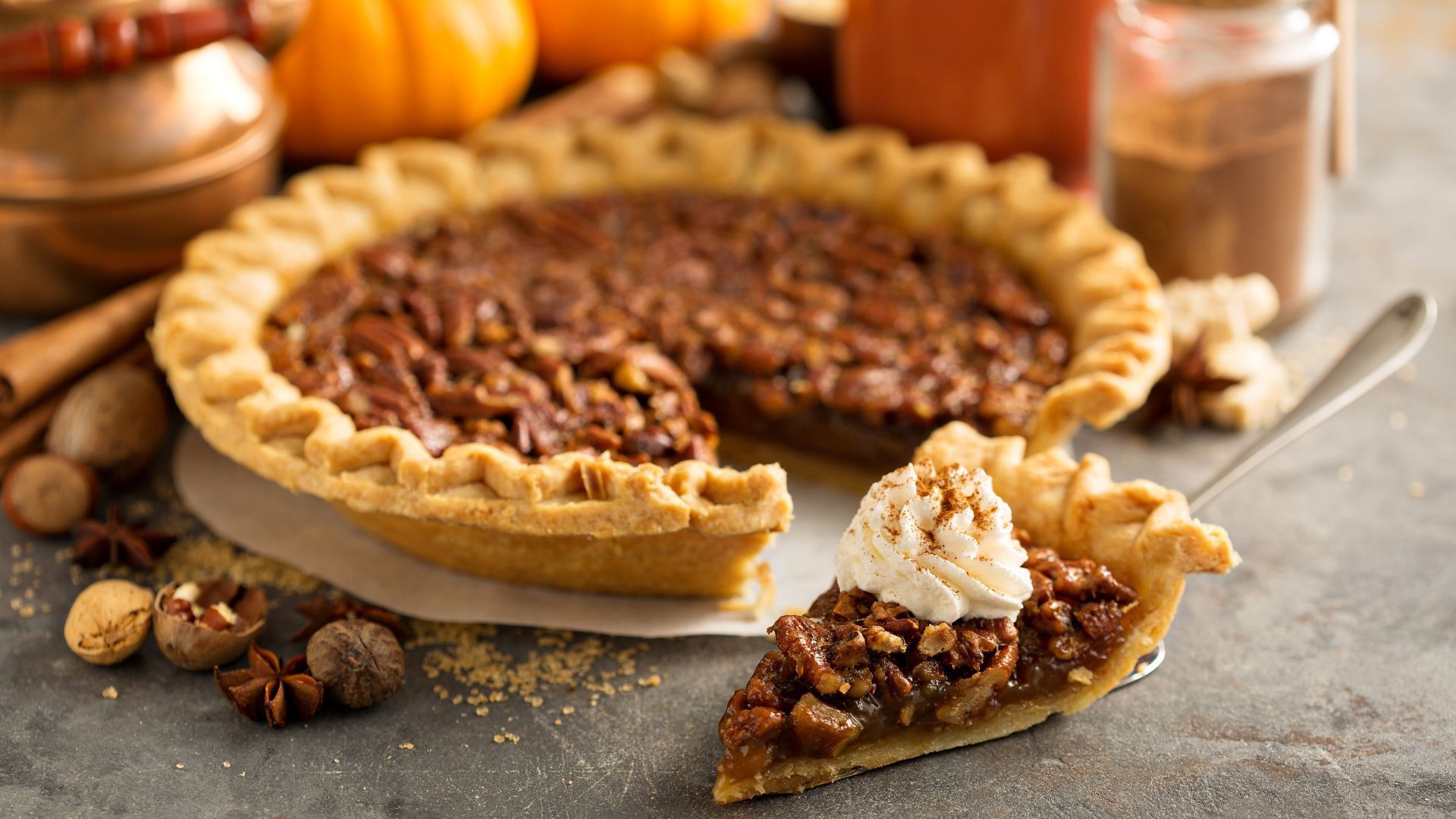 Pecan pie with a slice served, topped with whipped cream. Fall setting with pumpkins, spices, and copper.