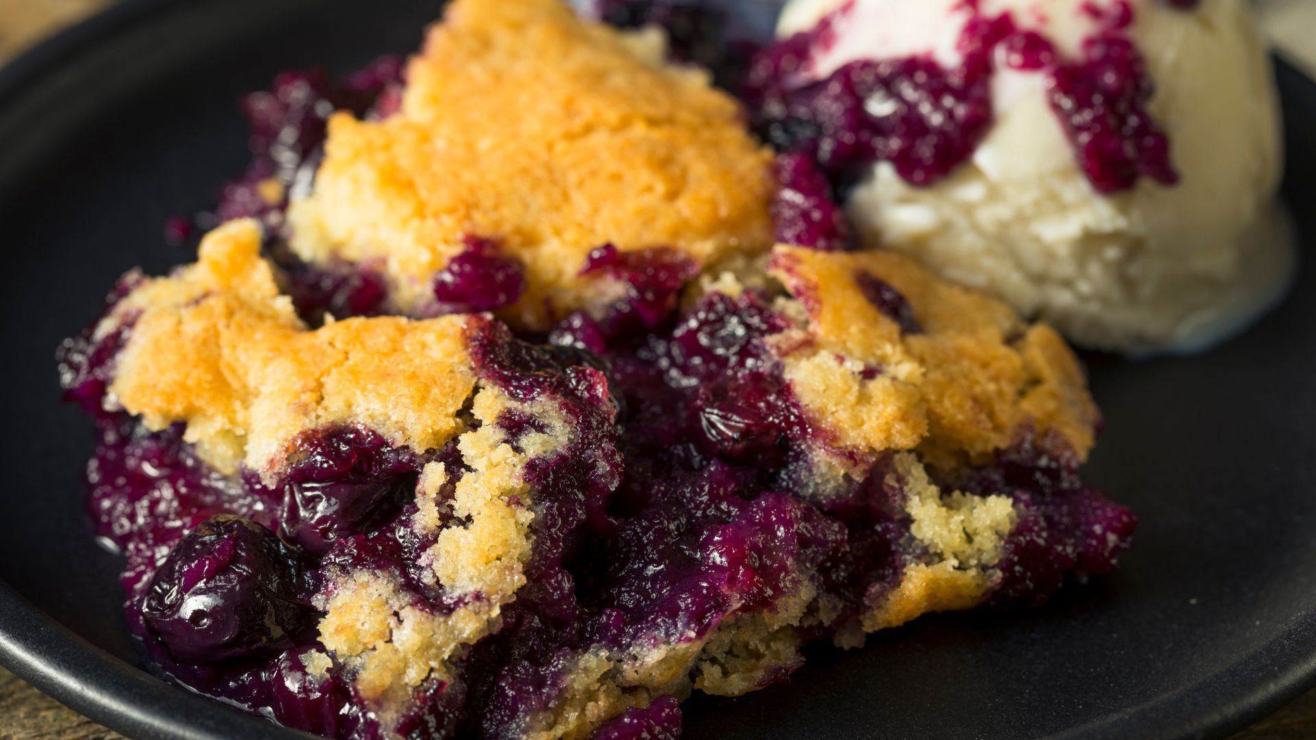 Blueberry cobbler with ice cream on a black plate.