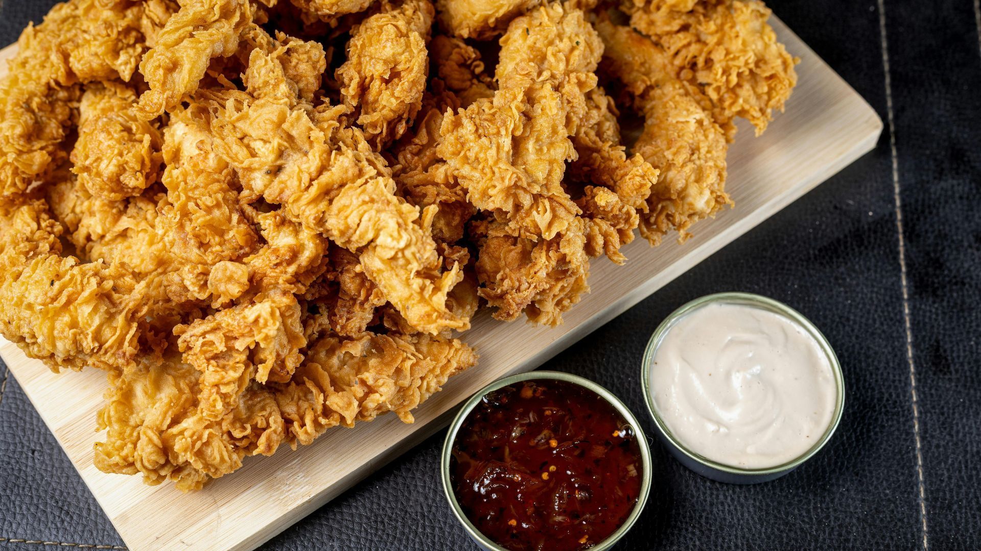 Fried chicken tenders on a wooden board with BBQ and white dipping sauces.