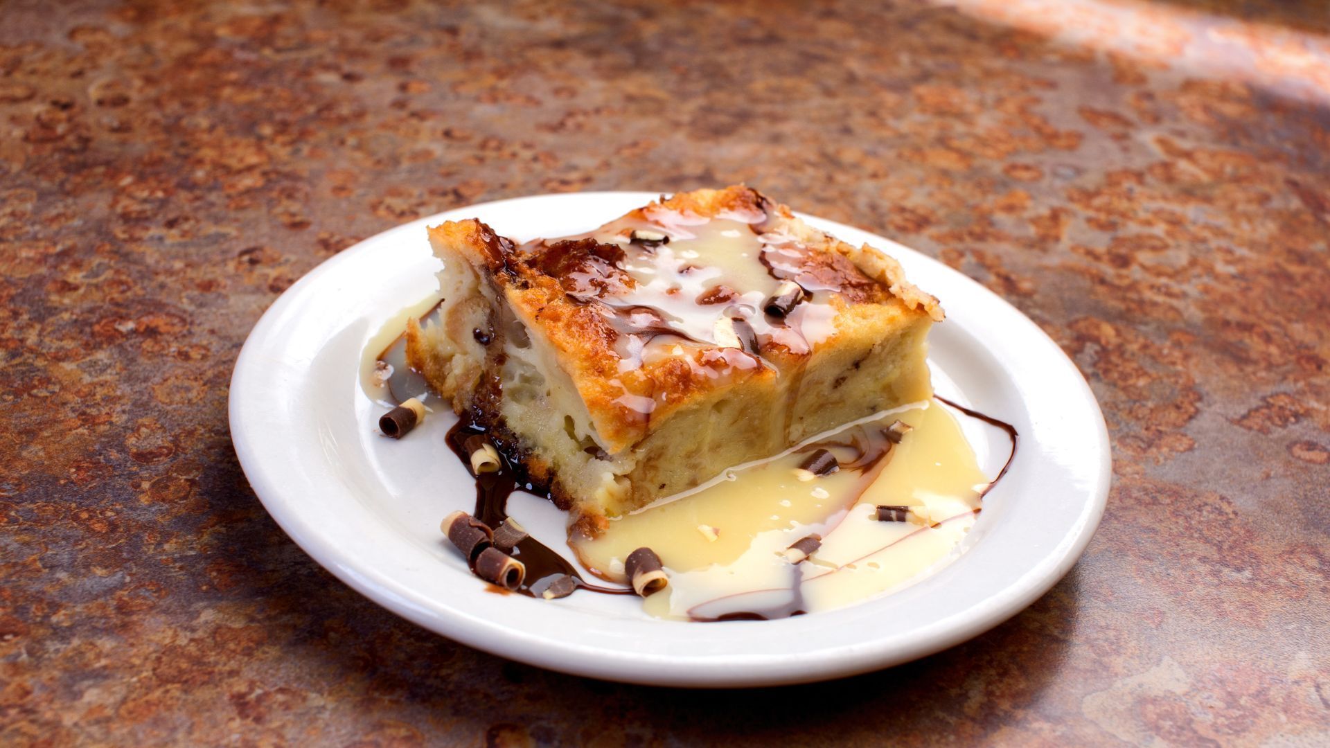 Bread pudding on white plate, drizzled with sauce and chocolate chips, on a brown surface.