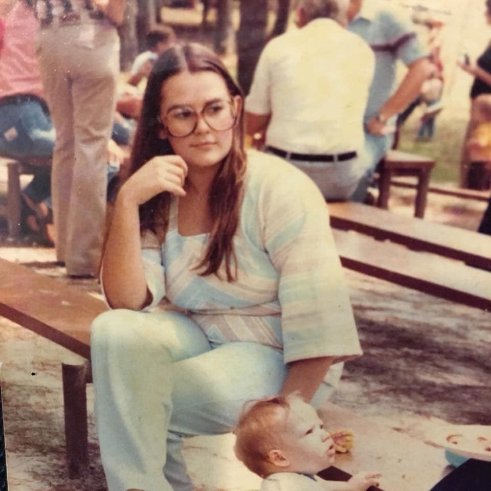 Woman wearing glasses sits on bench with a baby. People are in the background, outdoors.