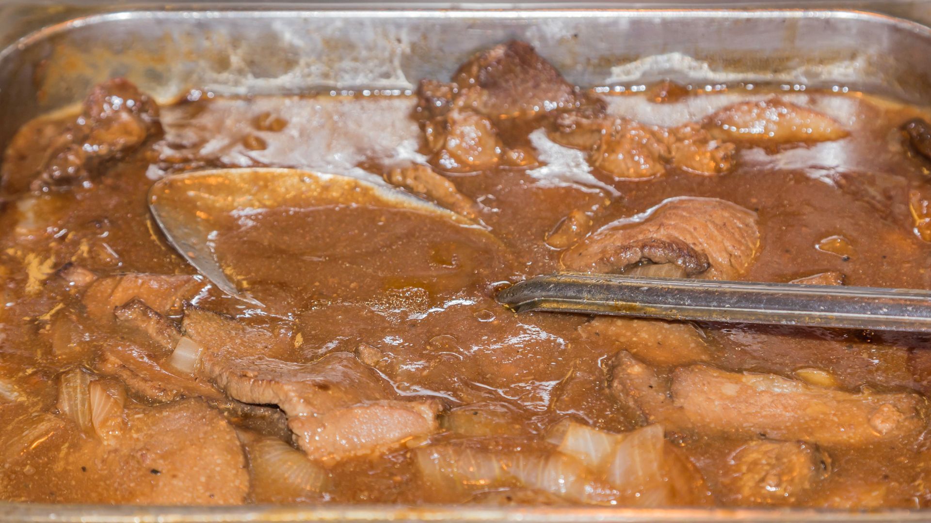 Meat in brown gravy in a metal pan, served with a ladle.