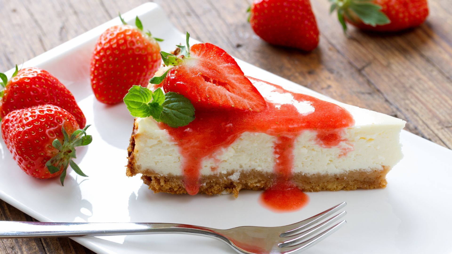 Slice of cheesecake with strawberry sauce and fresh strawberries on a white plate.