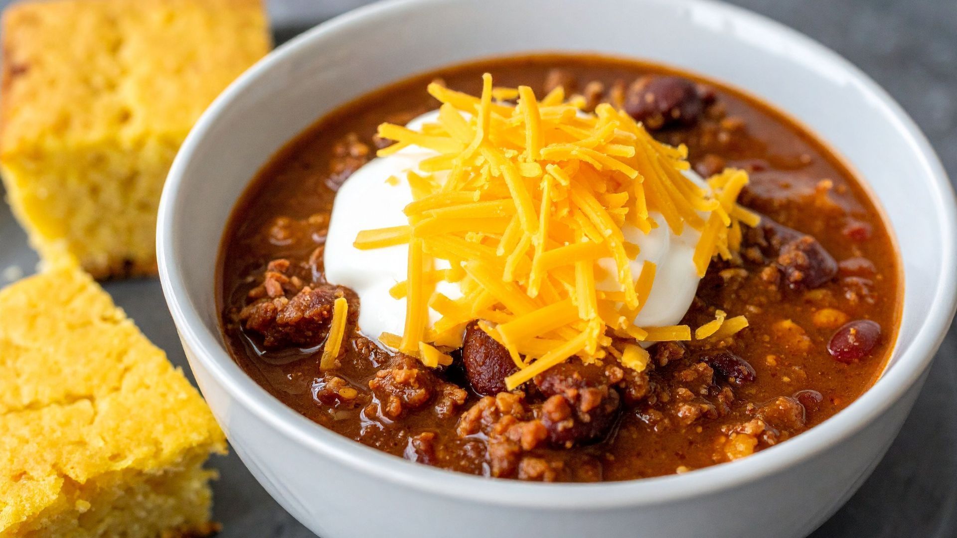 Bowl of chili topped with sour cream and shredded cheese, served with cornbread.
