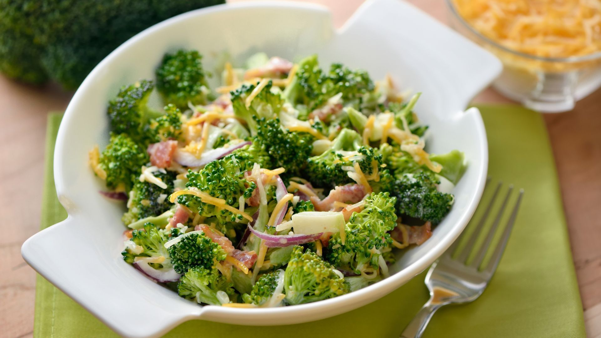 Broccoli salad in a white bowl, with shredded cheese, red onion, and bacon, on a green placemat.