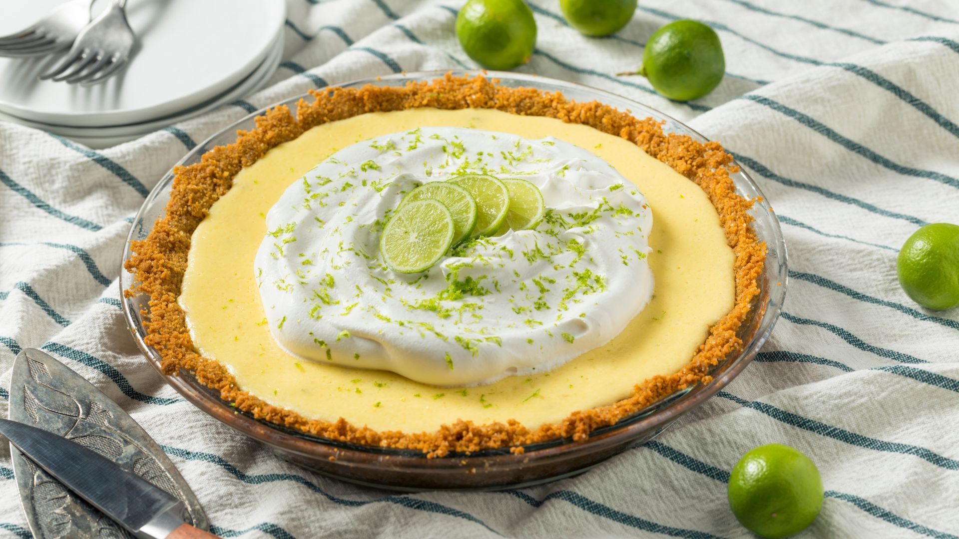 Key lime pie with whipped cream topping and lime slices, on striped cloth, with limes.