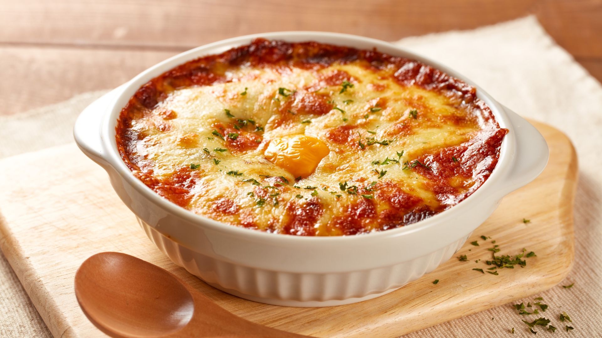 Baked dish with melted cheese in a white bowl, on a wooden cutting board with a spoon.
