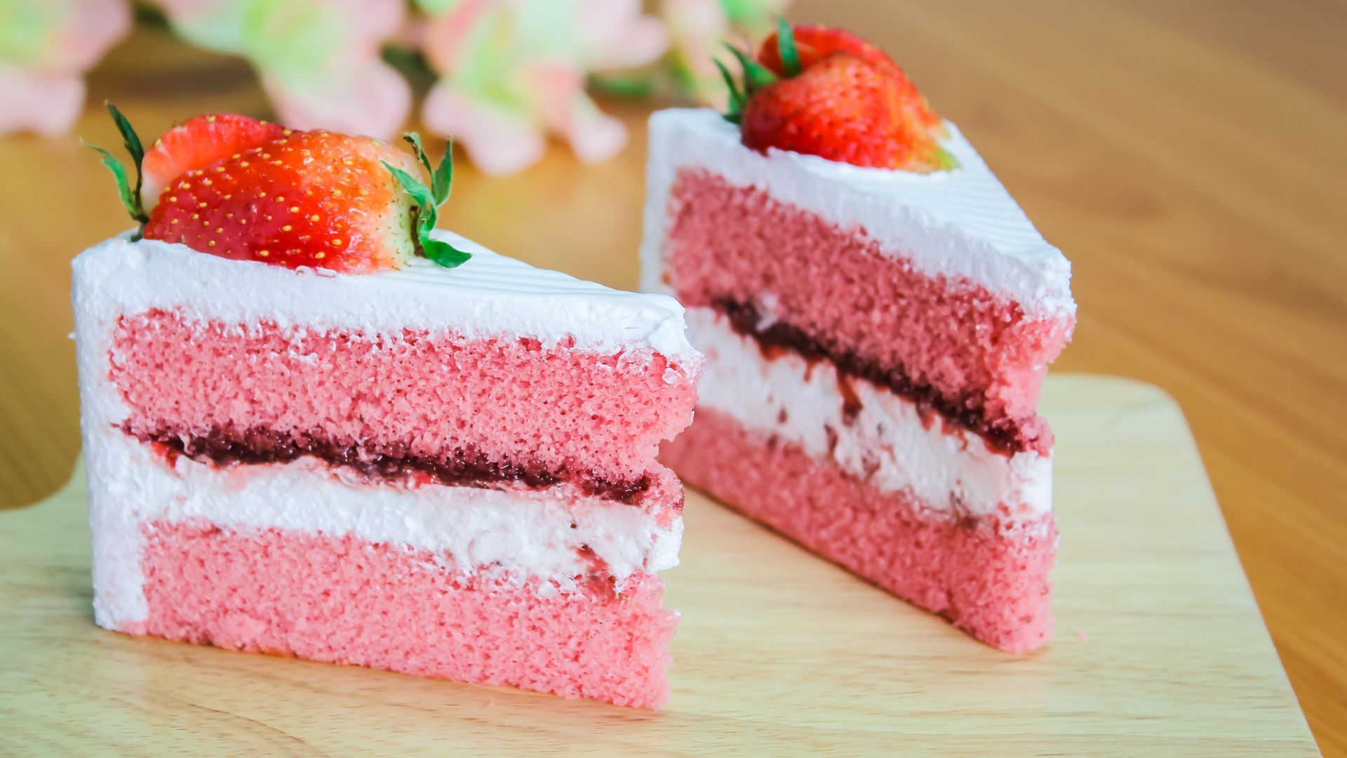 Two slices of pink cake with white frosting and strawberry topping on a wooden board.