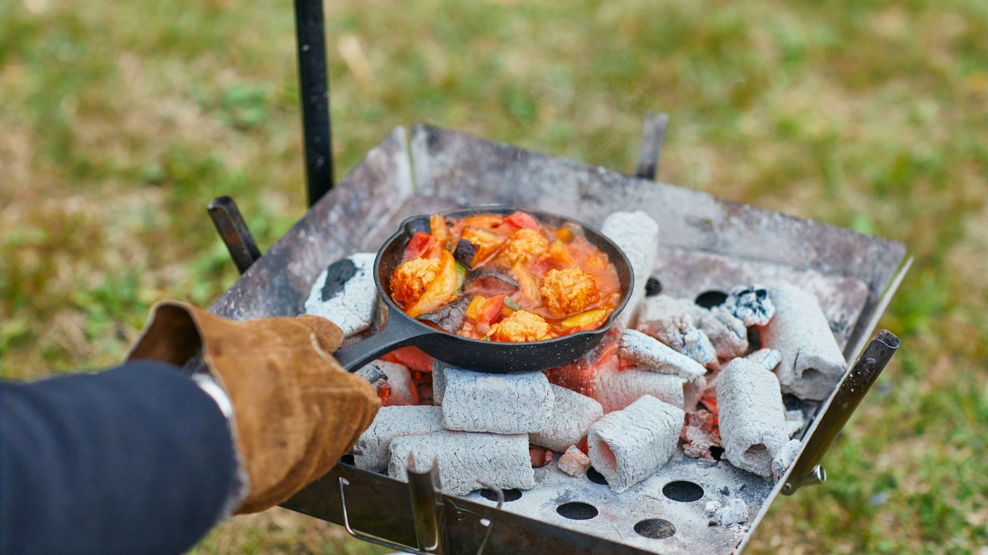 Person grilling food in a small cast iron pan over hot coals outdoors.